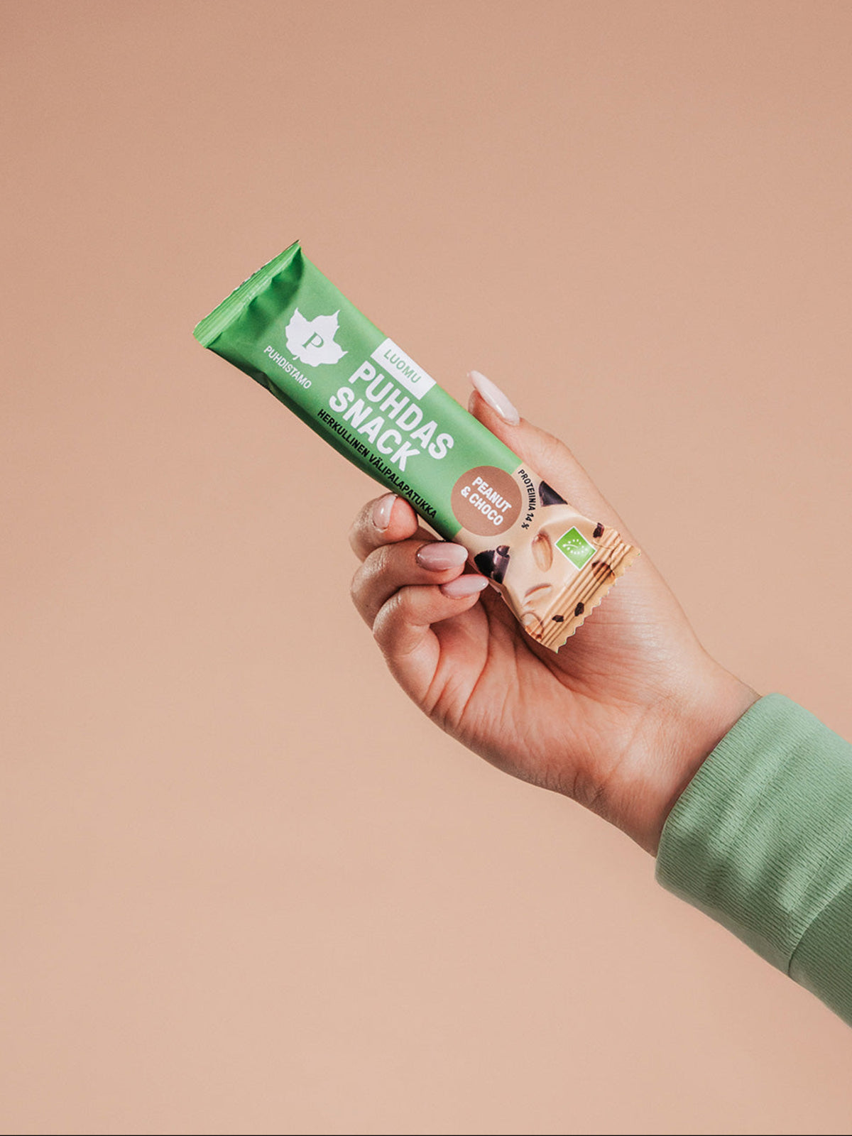 Nainen pitelee Puhdistamon Puhdas Snack Välipalapatukkaa kädessä beigeä taustaa vasten