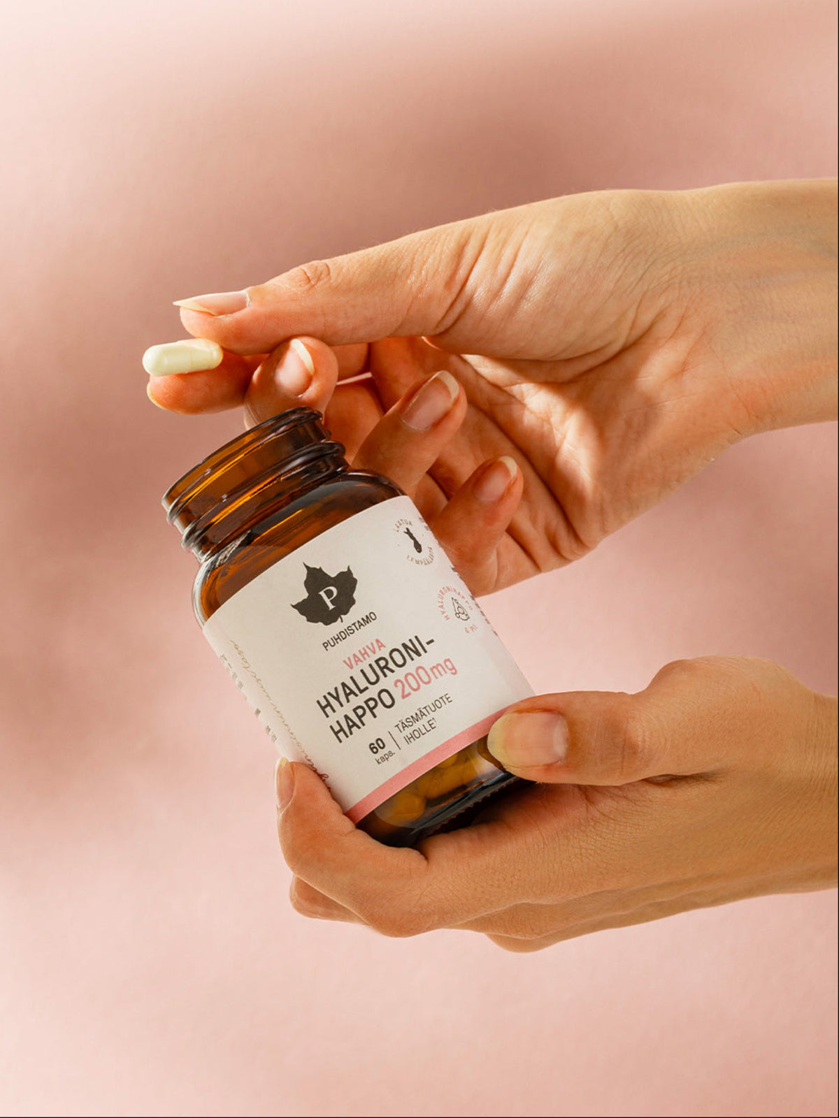 Nainen pitää kädessään Puhdistamon Vahva Hyaluronihappo 60 kapselin purkkia vaaleanpunertavaa taustaa vasten