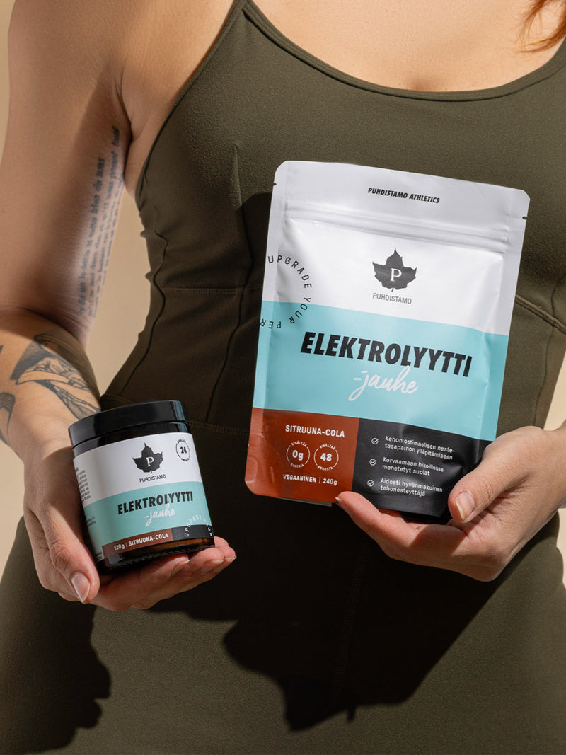 Nainen pitelee Puhdistamon Elektrolyyttijauhepurkkia ja -pussia maussa Sitruuna-Cola